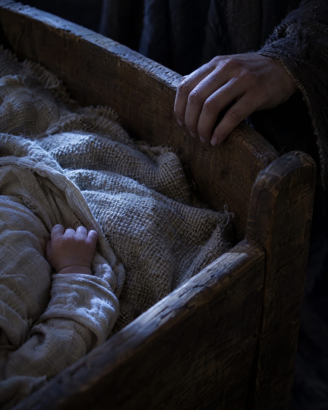 A mother's hand rests on the edge of a wooden cradle at night, beside her baby's small hand on the wool blanket.